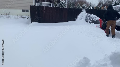 Rear three-quarter view of an adult man dressed in a casual gray hoodie, khaki pants with a visible cellphone in the rear pocket, and sturdy boots as he pushes a gasoline-powered snow blower, ejecting
