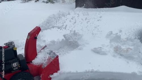 Side view of a casual adult man wearing a hooded sweatshirt, khaki jeans with a cellphone visible in his back pocket, and winter boots as he operates a push-style gasoline-powered snow blower.