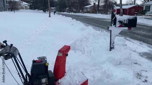 Side view of an adult man wearing a casual hooded sweatshirt and winter boots as he pushes a gasoline-powered snow blower, discharging snow to the side while clearing a residential driveway. no gloves