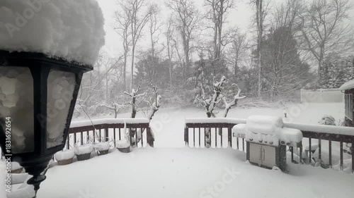 Video snapshot of a suburban backyard deck blanketed in fresh snow at night. A gas grill sits covered near the railing while a close-up outdoor lamp glows softly, illuminating falling snow and leafles