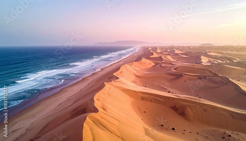 Fototapeta Naklejka Na Ścianę i Meble -  An aerial view of a coastline with sand dunes meeting the ocean at sunset. The image shows a vast expanse of sand dunes and the ocean with waves.