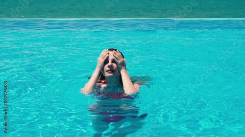 A girl traveler emerges from an infinity swimming pool with a beautiful ocean view at a luxury resort hotel villa. Drops of water run down her face and hair. Vacation travel tourism woman in bikini.