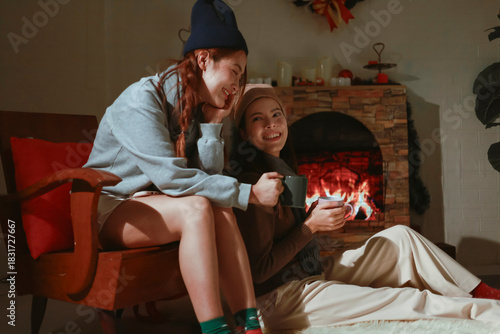 Teen girls having fun together on holiday, sitting in front of the fireplace. Asian lesbian couple celebrating Christmas, drinking warm drinks to keep warm.