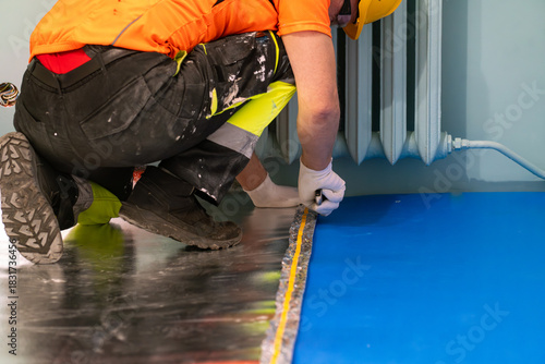 A finishing worker applies an insulating underlayment with an aluminum layer under wooden floor panels. Sound and thermal insulation. Apartment renovation.