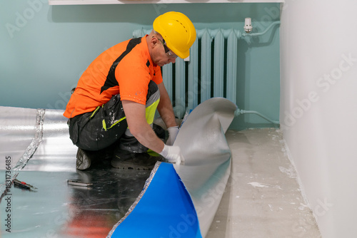 A contractor applies an insulating underlayment with an aluminum layer under wooden floor panels. Sound and thermal insulation. Apartment renovation.