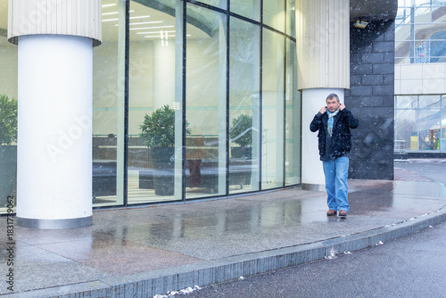 Man adjusting collar while moving along wet pavement near glass facade of contemporary building during light winter snowfall. High quality photo