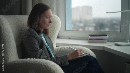 A woman sits comfortably in a soft chair, writing in a notebook. Sunlight pours in through the window, creating a warm atmosphere during the day.