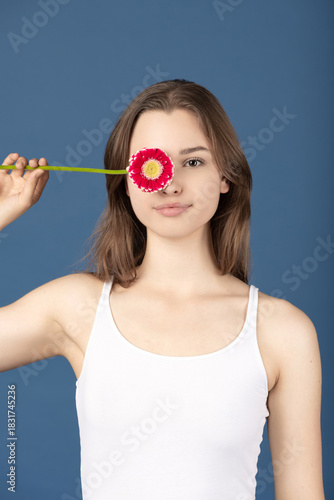 Beauty, make-up, style concept. Beautiful woman covering her eye with purple flower blossom. Model wearing white shirt and standing against blue background