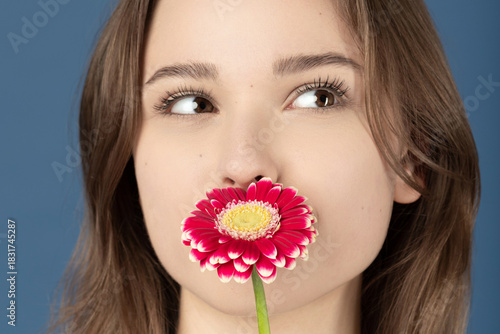 Beauty, make-up, style concept. Beautiful woman covering her mouth with purple flower blossom. Model wearing white shirt and standing against blue background