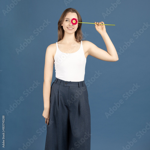 Beauty, make-up, style concept. Beautiful woman covering her eye with purple flower blossom. Model wearing white shirt and standing against blue background
