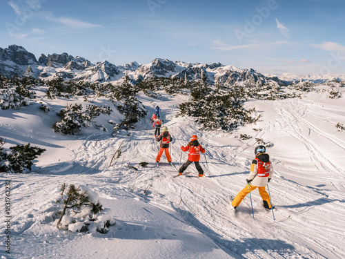 A group of children ski down a snowy slope, with snow-covered mountains in the background under a blue sky. Paganella, Roda refuge,Trentino, Italy