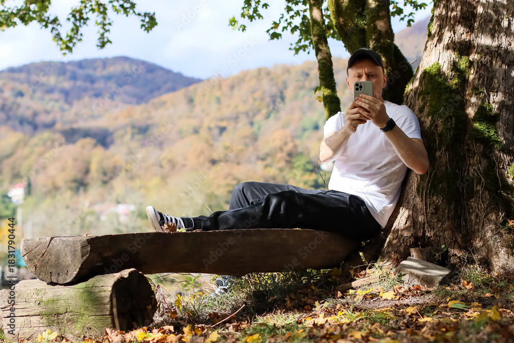 Fototapeta premium Seated male using phone camera for selfie in vibrant woodland with mountain horizon