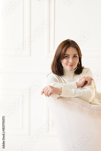 Woman in white sweater smiling while relaxing on a cozy chair in a bright, minimalist indoor space
