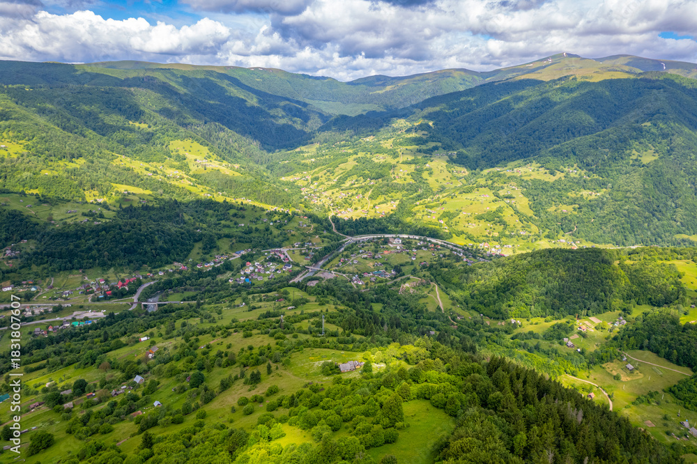 Obraz premium A high-altitude aerial view of a scenic mountain valley with lush green hills, forests, and scattered rural homes, showcasing a peaceful natural landscape under a dramatic cloudy sky.