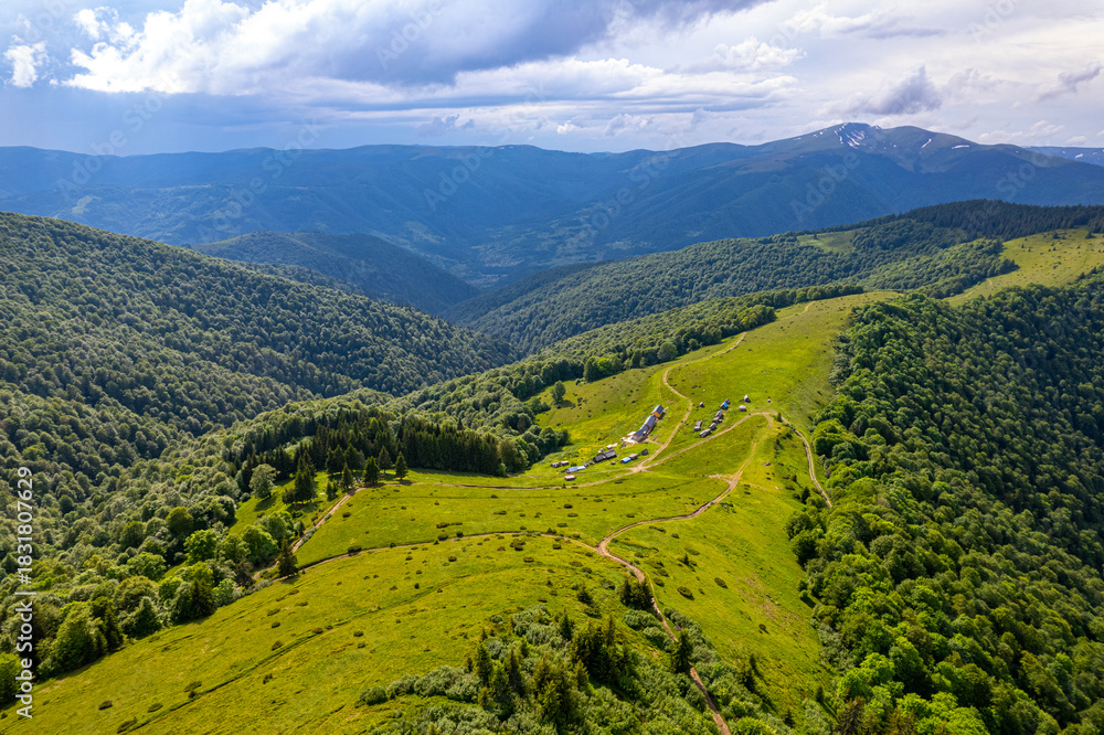 Fototapeta premium A high-altitude aerial view of a scenic mountain valley with lush green hills, forests, and scattered rural homes, showcasing a peaceful natural landscape under a dramatic cloudy sky.