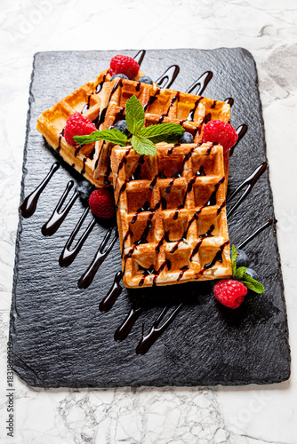 Sweet and crispy waffles sit on a dark slate, accented by fresh berries and chocolate sauce
