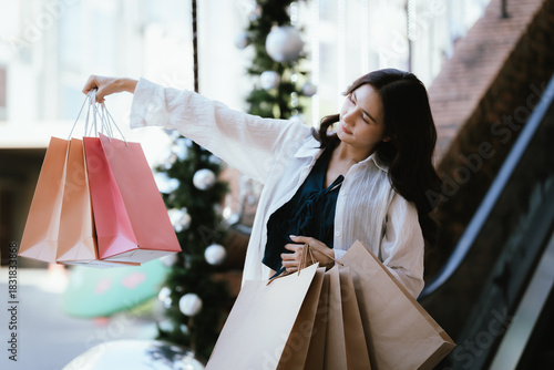 A smiling woman carries multiple shopping bags while walking outdoors near festive Christmas decorations, creating a cheerful holiday shopping vibe.