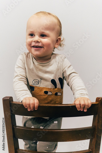 Portrait of a one-year-old child with beautiful curly hair standing on a chair by a white wall and smiling brightly, creating a warm and joyful atmosphere.