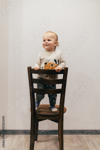 Portrait of a one-year-old child with beautiful curly hair standing on a chair by a white wall and smiling brightly, creating a warm and joyful atmosphere.