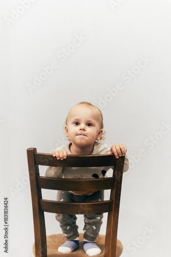 Portrait of a one-year-old child with beautiful curly hair standing on a chair by a white wall and smiling brightly, creating a warm and joyful atmosphere.