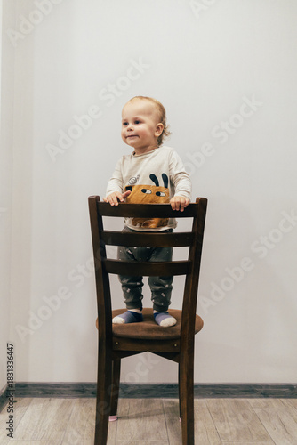 Portrait of a one-year-old child with beautiful curly hair standing on a chair by a white wall and smiling brightly, creating a warm and joyful atmosphere.