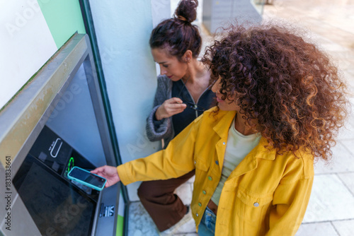 Εκτύπωση καμβά Young woman using smartphone for contactless payment at ATM with friend watching