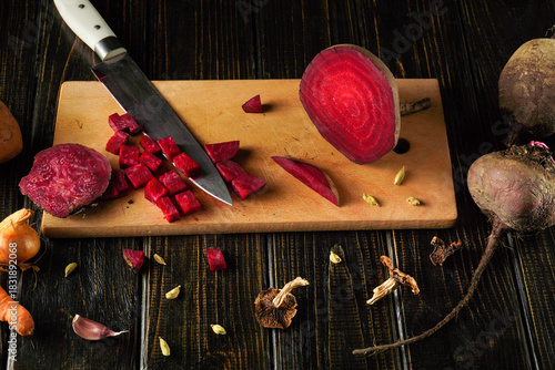 Fresh beets and various vegetables are being chopped on a wooden cutting board. A sharp knife is used to slice and prepare ingredients for a meal in a cozy kitchen