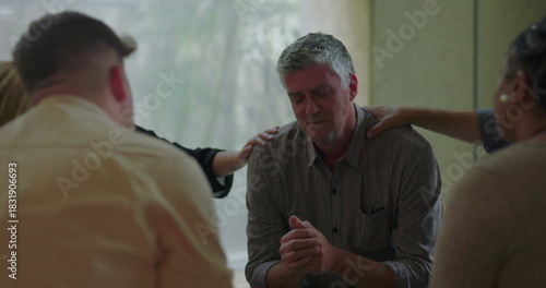 Fotografie Man bows head and clasps hands during group therapy session while surrounded by