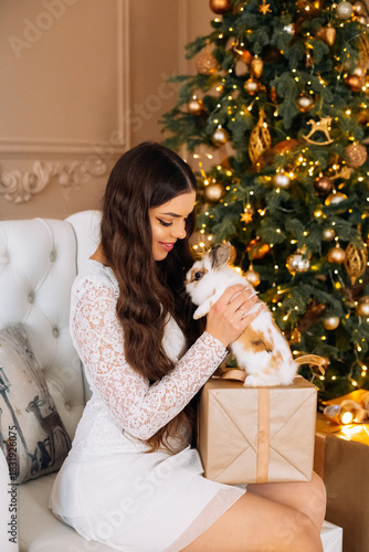 Woman in white dress smiles while holding a rabbit beside a beautifully decorated Christmas tree with warm lights and ornaments during the holiday season