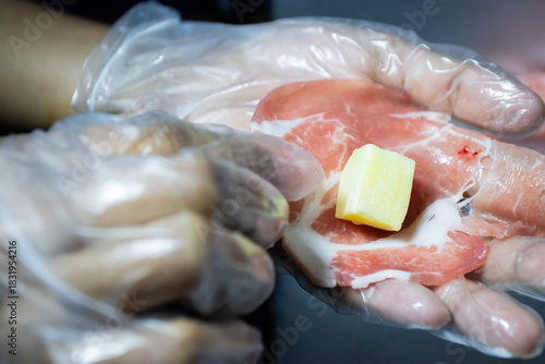 Gloved hand holding a piece of thinly sliced raw meat (pork or beef) with a small, yellow cube of cheese placed in the center, ready for preparation.