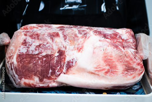 Large, frozen raw primal cut of red meat (pork or beef) with a thick layer of fat, held on a tray by a gloved hand, ready for slicing.