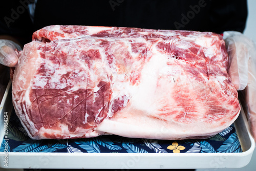 Large, frozen raw primal cut of red meat (pork or beef) with a thick layer of fat, held on a tray by a gloved hand, ready for slicing.