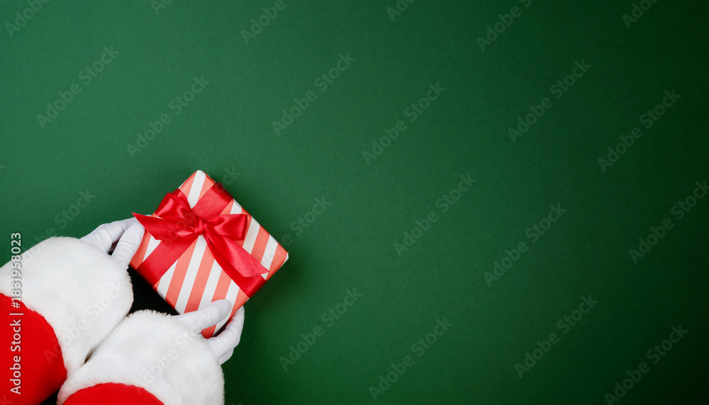Fototapeta premium Santa Claus hand holding red and white striped gift box with red ribbon on festive green background with copy space for text, Christmas card design element.