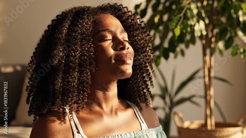 Young black woman with curly hair meditating or relaxing with eyes closed in warm sunlight near a houseplant indoors