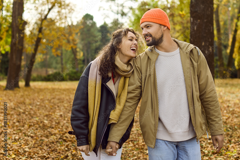 Fototapeta premium Couple in autumn park, smiling together. They hold hands among leaves, enjoying natures calm and romance outdoors laughing in cozy layers happy as they walk. Evokes love, joy and togetherness.