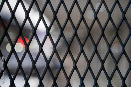 Close up view of dark metal expanded mesh or grating with a diamond-shaped pattern, with blurry lights and shapes in the background