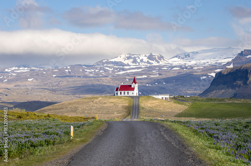 road leads to the small white concrete church with red roof with snow capped mountains in the background, Ingjaldshóll church, Snæfellsnes Peninsula, Iceland