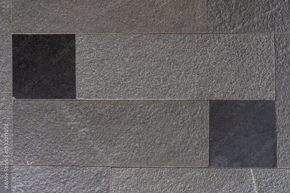 Fototapeta premium Textured wall surface composed of large grey rectangular natural stone tiles with rough surfaces and two smooth black inset stones