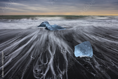 View of luminous ice diamonds scattered across the stark black sand beach as the tide ebbs and flows under a twilight sky, Diamond Beach, Sveitarfelagid Hornafjordur, Iceland.