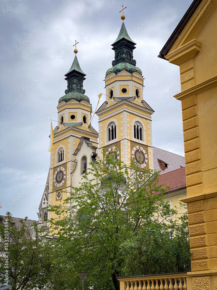 Naklejka premium Twin towers of Brixen Cathedral in South Tyrol, Italy, yellow facades with copper spires and clock faces under a sky with clouds