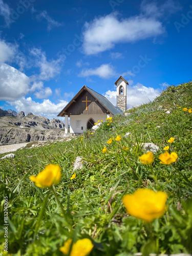 Low angle view of Cappella degli Alpini along Tre Cime hiking trail, Italy with blurred alpine meadow and a yellow buttercup (Ranunculus) in forerground against blue sky with clouds