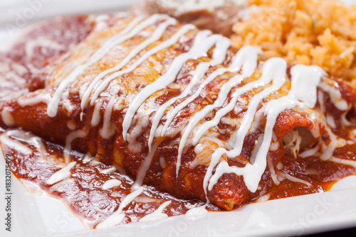 A plate of enchiladas with red Ranchero sauce, white cream and melted cheese with beans and rice.