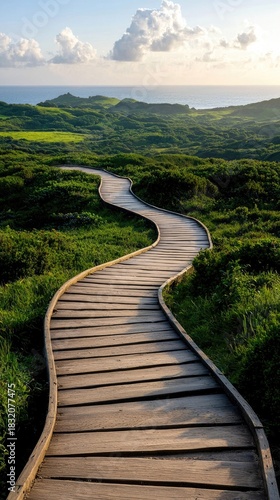 A wooden pathway curves through a vibrant green landscape, leading towards the ocean under a bright, sunny sky. The scene is filled with lush vegetation and rol