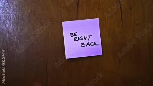 A small pink sticky note is placed on a wooden door, reminding the person to be back soon. The note emphasizes a friendly and casual communication style.