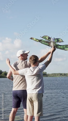 Father and son launching a plane-shaped kite together by the lake on Father's Day. Vertical shot video.