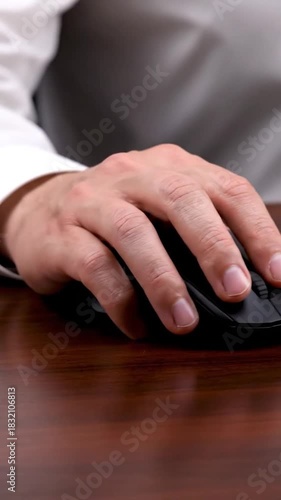 A hand on a black computer mouse resting on a wooden desk. Close up view