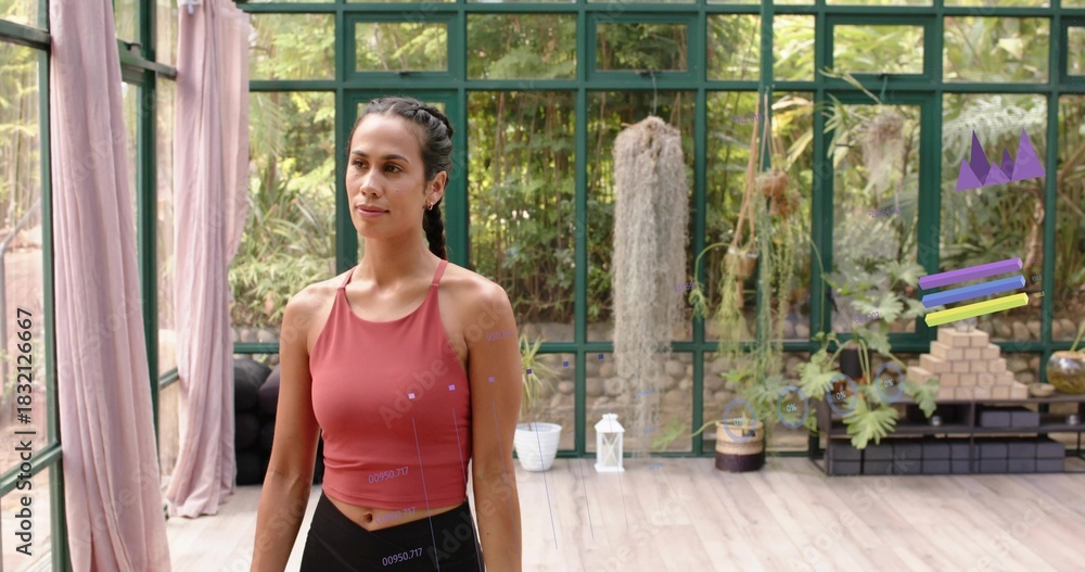 Fototapeta premium Standing Pacific-Islander woman in rust top and leggings stretching in glass studio with plants