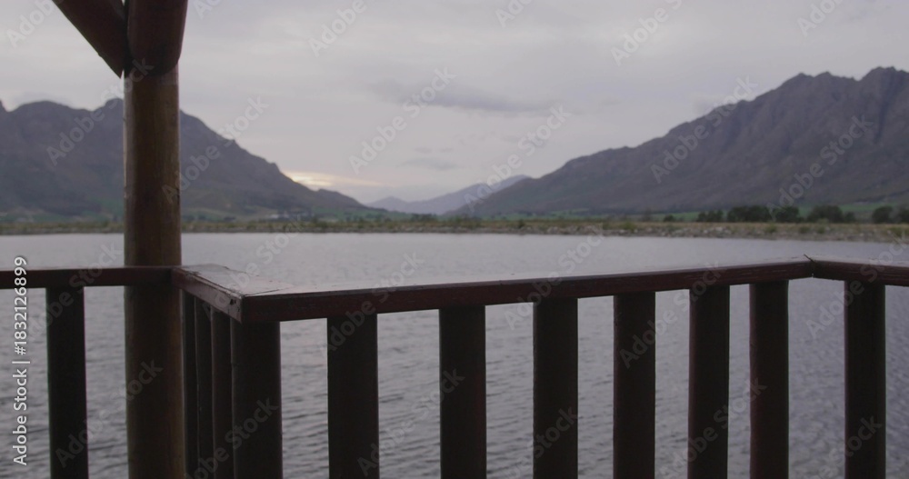 Fototapeta premium Framing wooden railing posts overlooking calm lake, showing rugged mountains and overcast sky