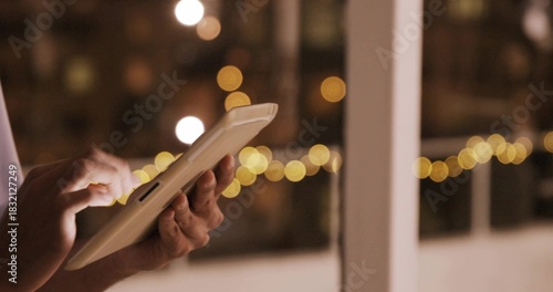 Tapping adult hands with light sleeve cuff using white tablet screen near glass door, string lights