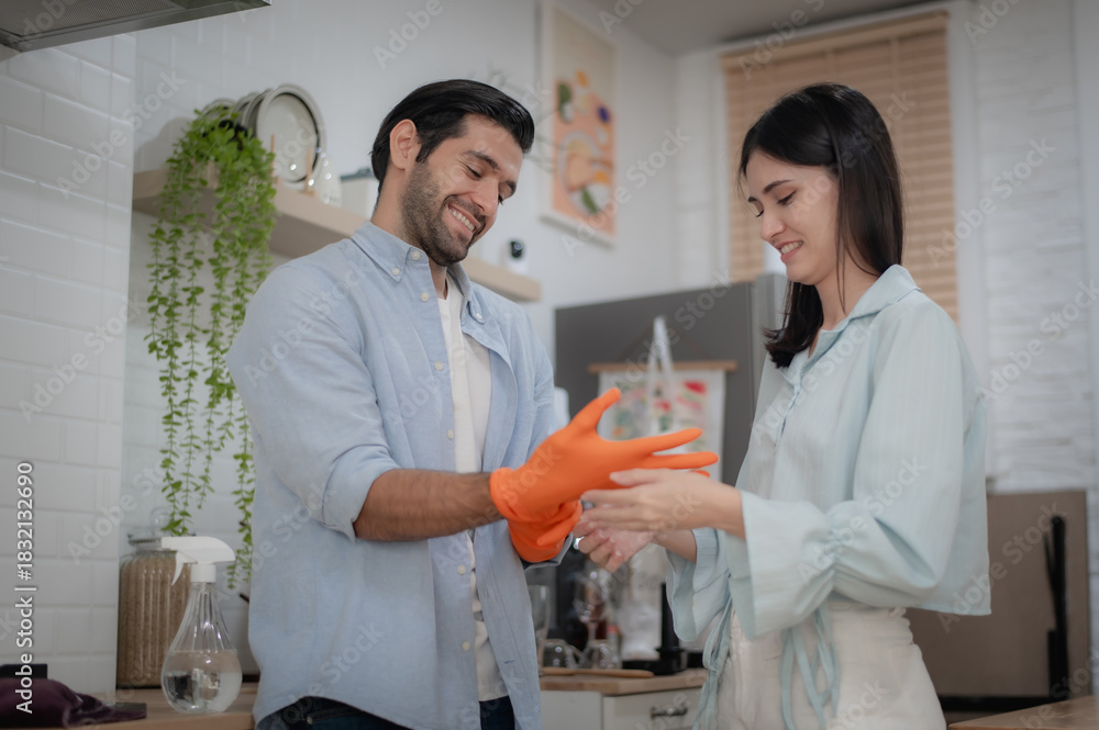 Fototapeta premium Couple preparing to clean kitchen, helping each other put on gloves.